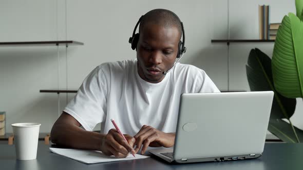 Focused Black Businessman Wearing Headphones Writing Notes in a Notebook Watching a Video Course