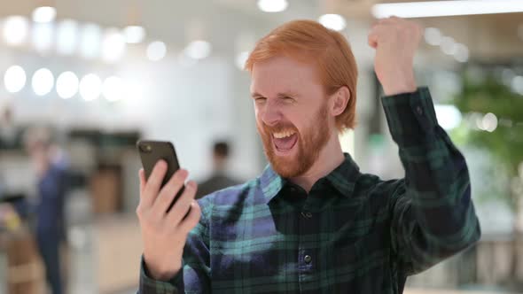 Portrait of Beard Redhead Man Celebrating Success on Smartphone 