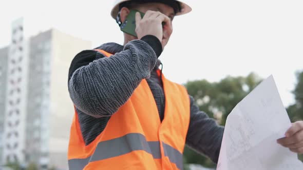 The foreman at the construction site is talking on the phone and checking the drawings.