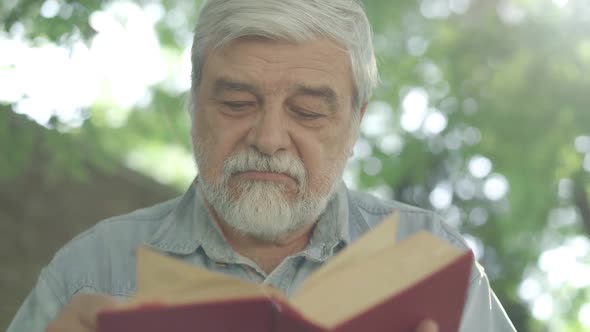 Close-up Face of Grey-haired Bearded Old Man with Brown Eyes Reading Book in Sunlight Outdoors