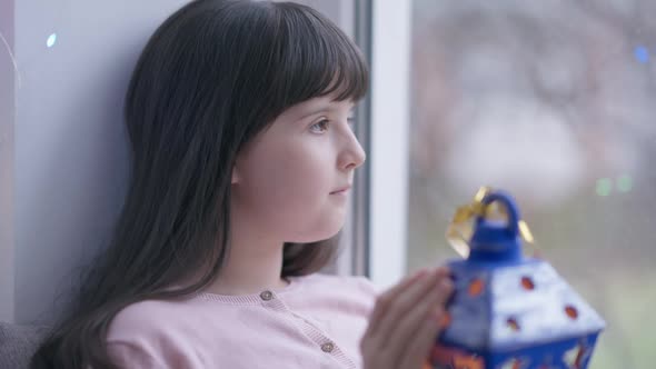 Closeup Portrait Caucasian Girl with Christmas Lantern Looking Out the Window Sitting on Windowsill