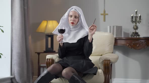 Wide Shot Portrait of Slim Gorgeous Caucasian Woman in Nun Costume Sitting Indoors with Wine Glass