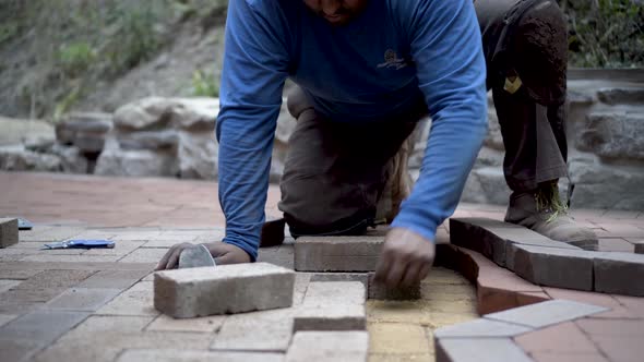 Very tight shot of stone mason fitting brick pavers into a herringbone patio design with an emblem i