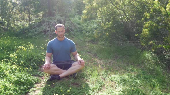 A man sitting in a meditation pose in a green sunny forest meadow practicing deep breathing exercise