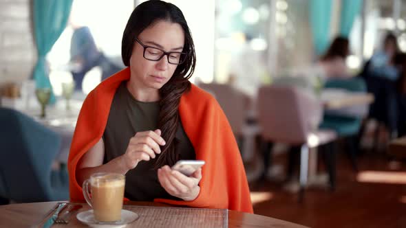 A Focused Woman with Glasses Writes a Message on Her Smartphone While Sitting at a Cafe Table with a