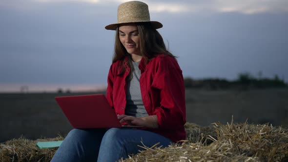 Happy Smiling Young Woman in Straw Hat Surfing Internet on Laptop Sitting in Twilight on Hay Bale