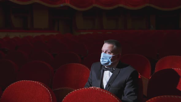 Sad man in medical mask in empty theater. Rich man in suit sitting alone among empty red armchairs