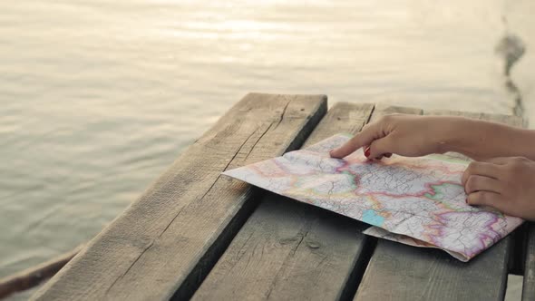 Woman Chooses the Route of Her Trip at Map on Sea Pier at Sunset, Hands Closeup