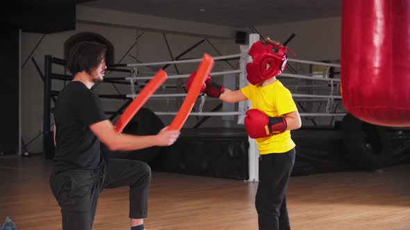 Little Boy in Protective Helmet Doing Boxing with a Young Coach  Hitting in the Long Soft Sticks