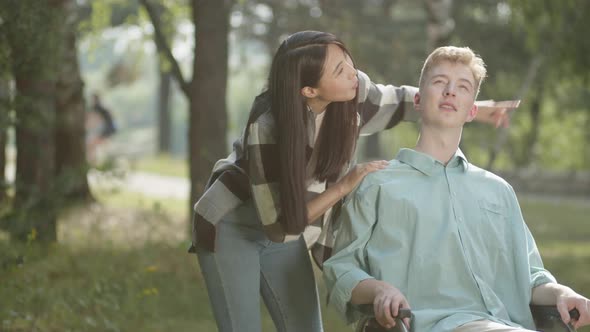 A Cute Asian Lady with Long Dark Hair Shows Her Disabled Young Man in a Blue Shirt Something in the