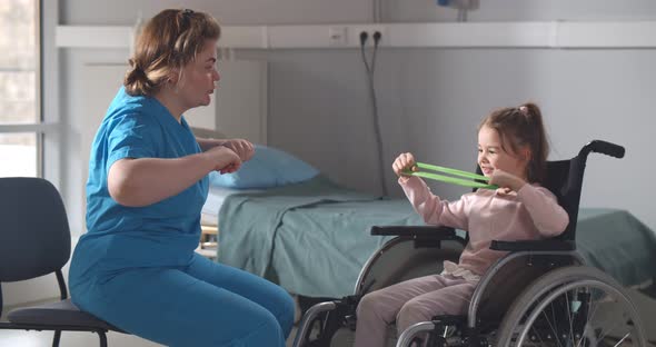 Nurse Helping Little Girl Patient in Wheelchair During Physical Exercise with Resistance Band