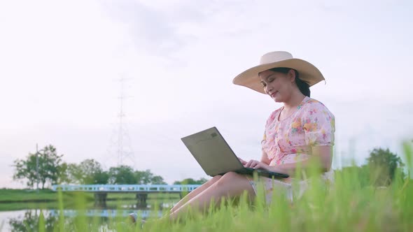 Beautiful Asian woman in hat sitting on lawn working on computer feeling successful.