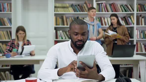 Bearded Modern Black-Skinned Guy which Sitting in the Library and Using Tablet PC