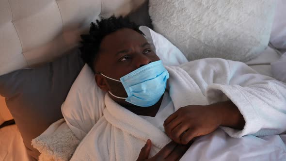 Closeup Portrait of Ill African American Young Man Coughing Lying in Bed in Coronavirus Face Mask