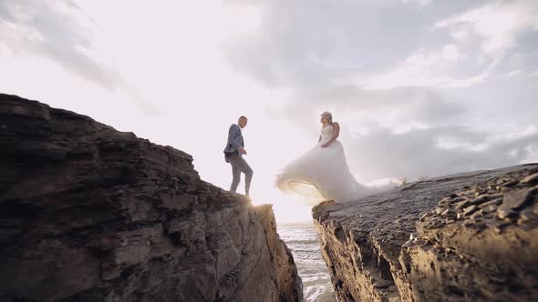 Groom Goes To Bride and Gives Her a Hand. Newlyweds on Mountainside By the Sea