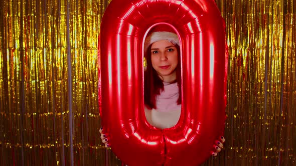 Woman in Santa Hat Holds Red Figure Zero on Background of Shimmering Golden Tinsel