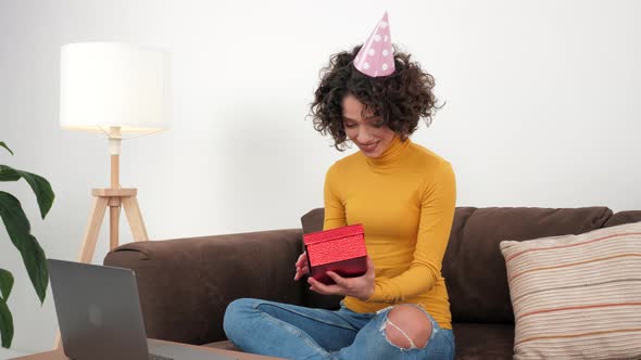 Happy Woman in Party Hat Opens Gift Box and Surprised Sitting in Couch at Home