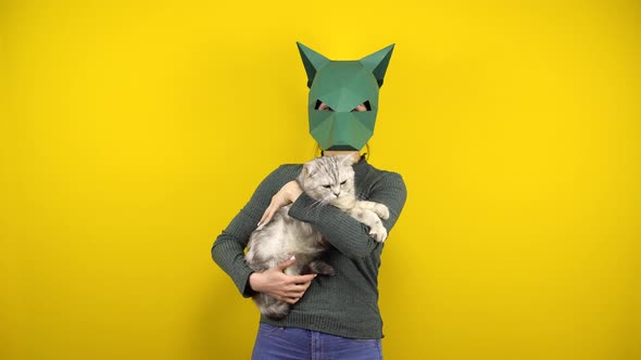 A Young Woman in a Cardboard Jackal Mask Holds a Cat in Her Arms on a Yellow Background. Woman in