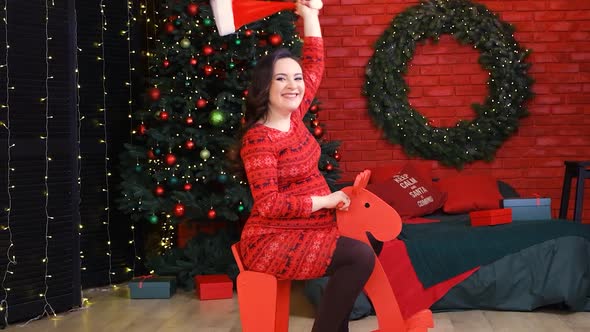 A Happy Pregnant Woman Rides a Red Wooden Toy Horse and Has Fun Preparing for Christmas