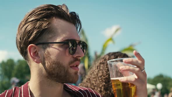 Close up of young caucasian man drinking beer from disposable cup at music festival and looking arou