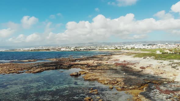 Drone view of sea coast with waves hitting rocky beach. Mountains and city buildings in Background. 