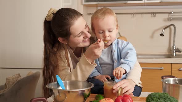 Cute Baby Boy Sitting on Kitchen Table and Playing with Cookware While Mother Feeding Him with