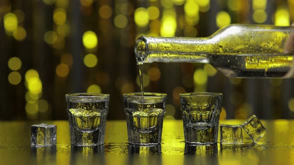 Barman Pour Frozen Vodka From Bottle Into Shot Glass. Ice Cubes Against Shiny Gold Party Background