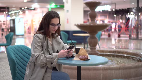 A Young Woman in a Cafe with a Phone in Her Hands Against the Backdrop of a Fountain