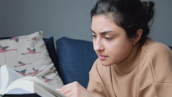 Close Up of a Young Indian Woman Sitting Next to a Sofa Thoughtfully Reading a Book