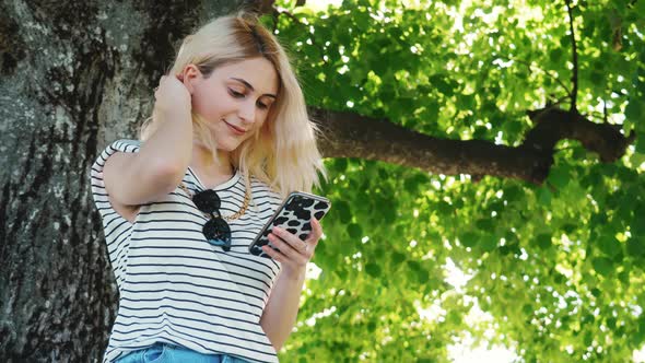 Young Cheerful Girl Sitting Close to a Tree and Reading Something Pleasant in Her Phone