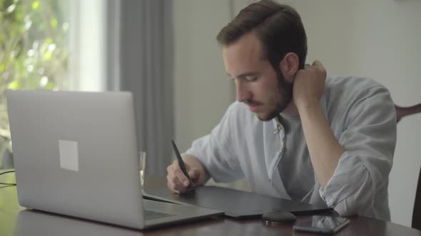 Successful Bearded Guy Draws Using Graphic Tablet Looking Into a Laptop