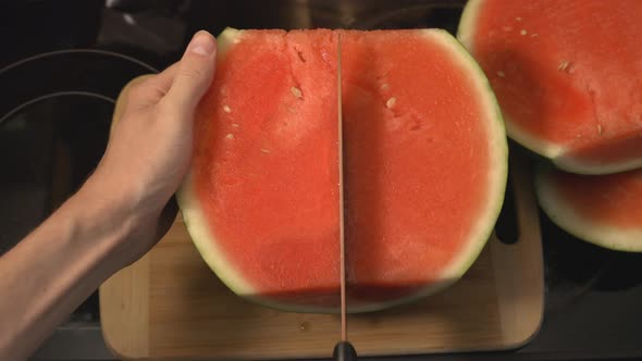 Hand Firmly Holding A Half-Sliced Watermelon Side Down Slicing In Half Lengthwise With A Sharp Knife