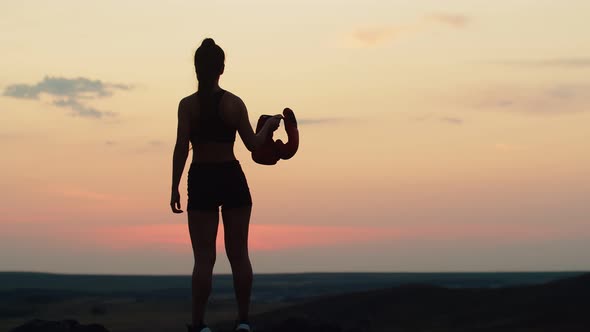 Female Boxer in Black Sports Shorts and Top Stands on Hill Against Sunset and Holds Boxing Gloves in