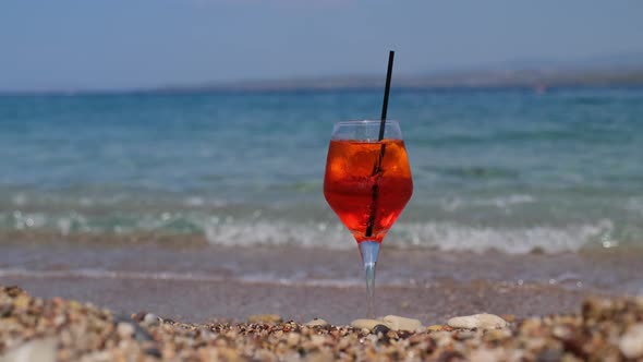 Glass of Aperol Spritz Cocktail Stands on Sand Near the Sea