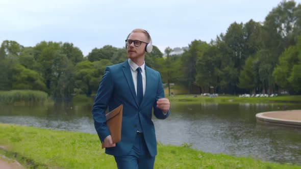 Excited Young Businessman Listening To Music in Headphones Walking in Summer Park