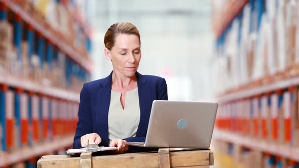 Female warehouse manager writing on clipboard and using laptop