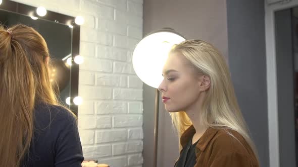 Young Stylist Putting Makeup on Face of Beautiful Blonde Girl