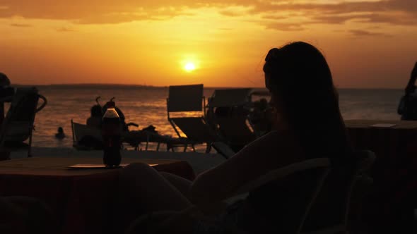 Silhouette Young Woman Sitting at Table on Beach and Looking at Sunset By Ocean