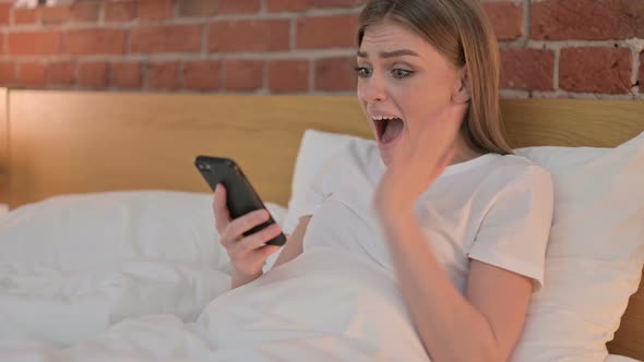 Cheerful Young Woman Celebrating Success on Smartphone in Bed 