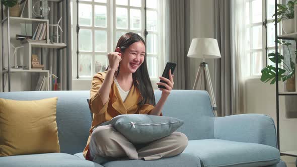 Asian Woman Sitting On Couch In Living Room Celebrating While Using Smartphone