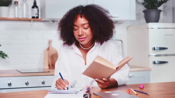 Young African American Woman Student Making Notes in Workbook Sits in Kitchen