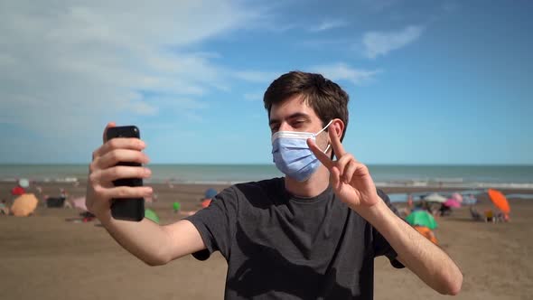 Young Man In Face Mask Taking A Selfie Showing Peace Sign At The Beach At Summertime]. - medium shot