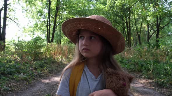 Little Girl Tourist in a Hat with Backpack and Teddy Bear Walks Alone in the Woods on Sunny Day