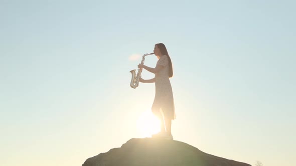 Beautiful Woman in Light Wind Blown Dress Plays Saxophone Against Backdrop of Sunset