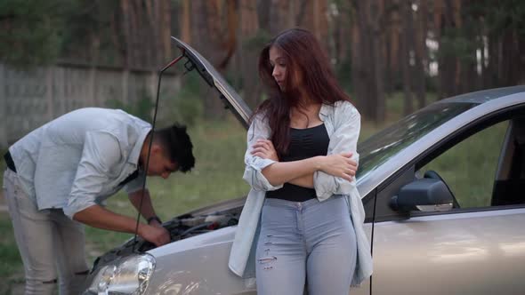 Bored Dissatisfied Girlfriend Leaning on Car in Forest Talking to Man Repairing Broken Vehicle