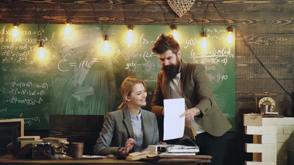 Professor Assisting Young Woman Hold Book Against Chalkboard in Classroom. Woman Assistant