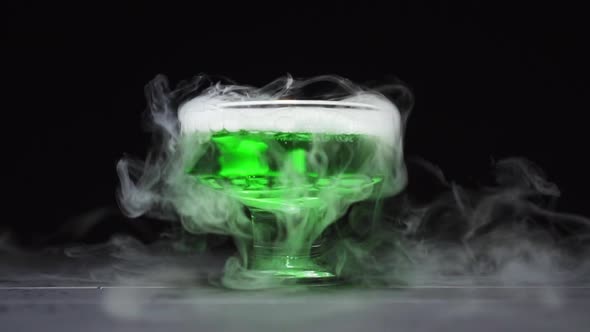 Green Liquid Boils Giving Off Smoke in a Glass Bowl on a Black Background