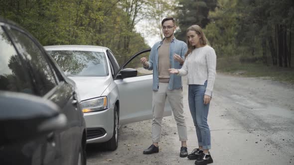 Wide Shot of Young Caucasian Man and Woman Talking and Gesturing Looking at Broken Cars on Roadside