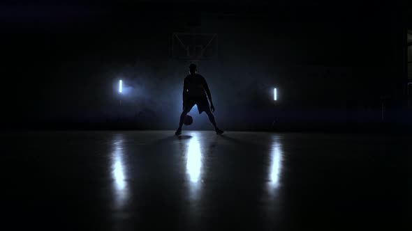 A Man with a Basketball on a Dark Basketball Court Against the Backdrop of a Basketball Ring in the