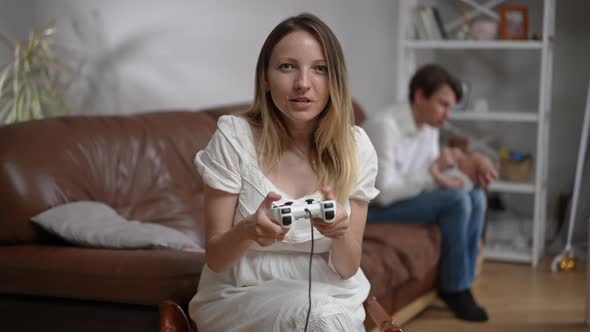 Carefree Young Mother Gaming Online with Joystick As Blurred Father Taking Care of Newborn Infant at